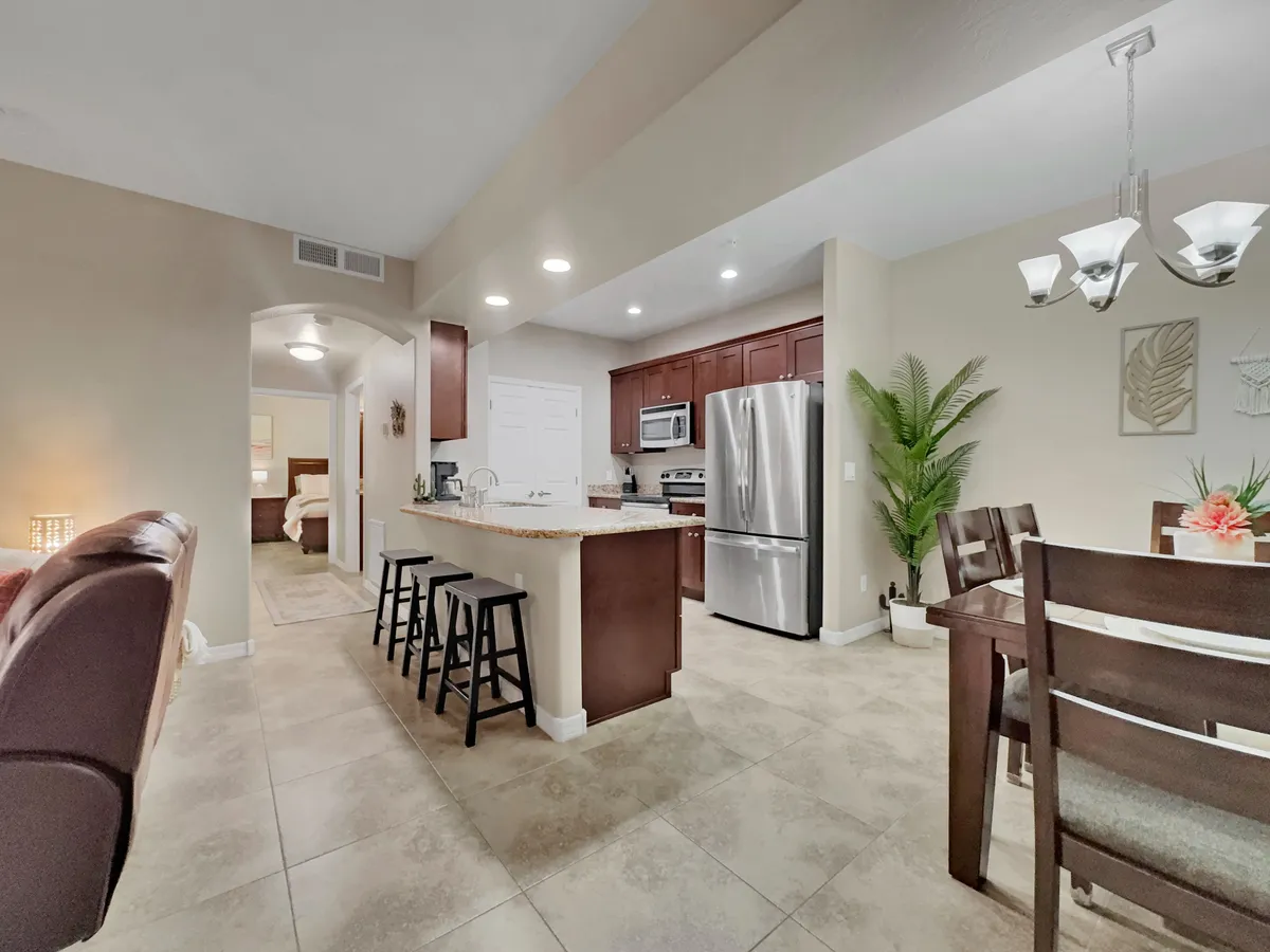 The image features a spacious kitchen island and adjacent dining table with stools, a refrigerator, and a potted plant, creating a hub for cooking, dining, and socializing. The scene is bright and inviting, ideal for group meals or celebrations.