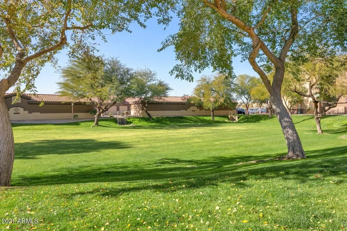 An inviting exterior scene featuring a lush green lawn dotted with mature trees, set against background residential buildings. The open space is ideal for outdoor games, picnics, and relaxed family gatherings under the shade.