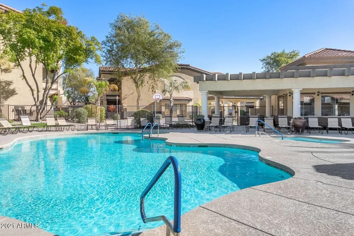 A vibrant exterior shot of a swimming pool surrounded by lush greenery, with poolside lounge chairs, a basketball hoop, and a covered seating area. The atmosphere is lively yet relaxed, suggesting opportunities for both active play and restful lounging.