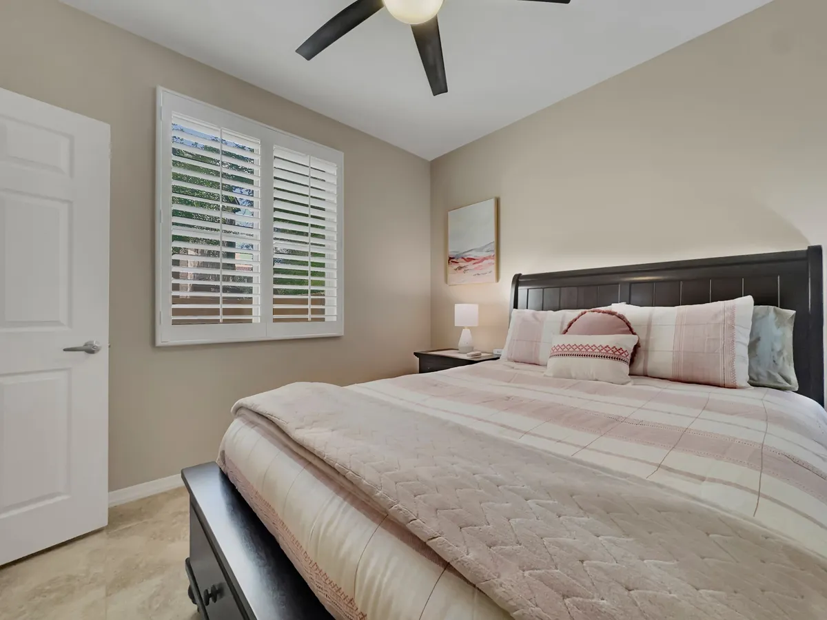 A tranquil bedroom featuring a cozy bed adorned with decorative pillows, wall art above the headboard, and a window with shutters letting in natural light. The space radiates warmth and comfort, ideal for a personal sanctuary.