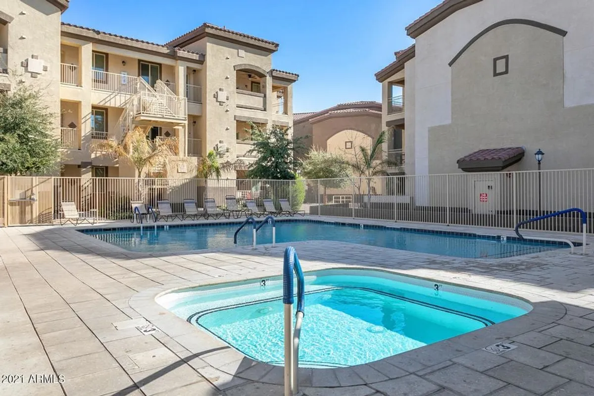 A striking exterior view of the resort-style swimming pool and jacuzzi surrounded by lush greenery and attractive building architecture. The pool area is inviting, with ample space for lounging, socializing, and relaxing in the sun or shade.