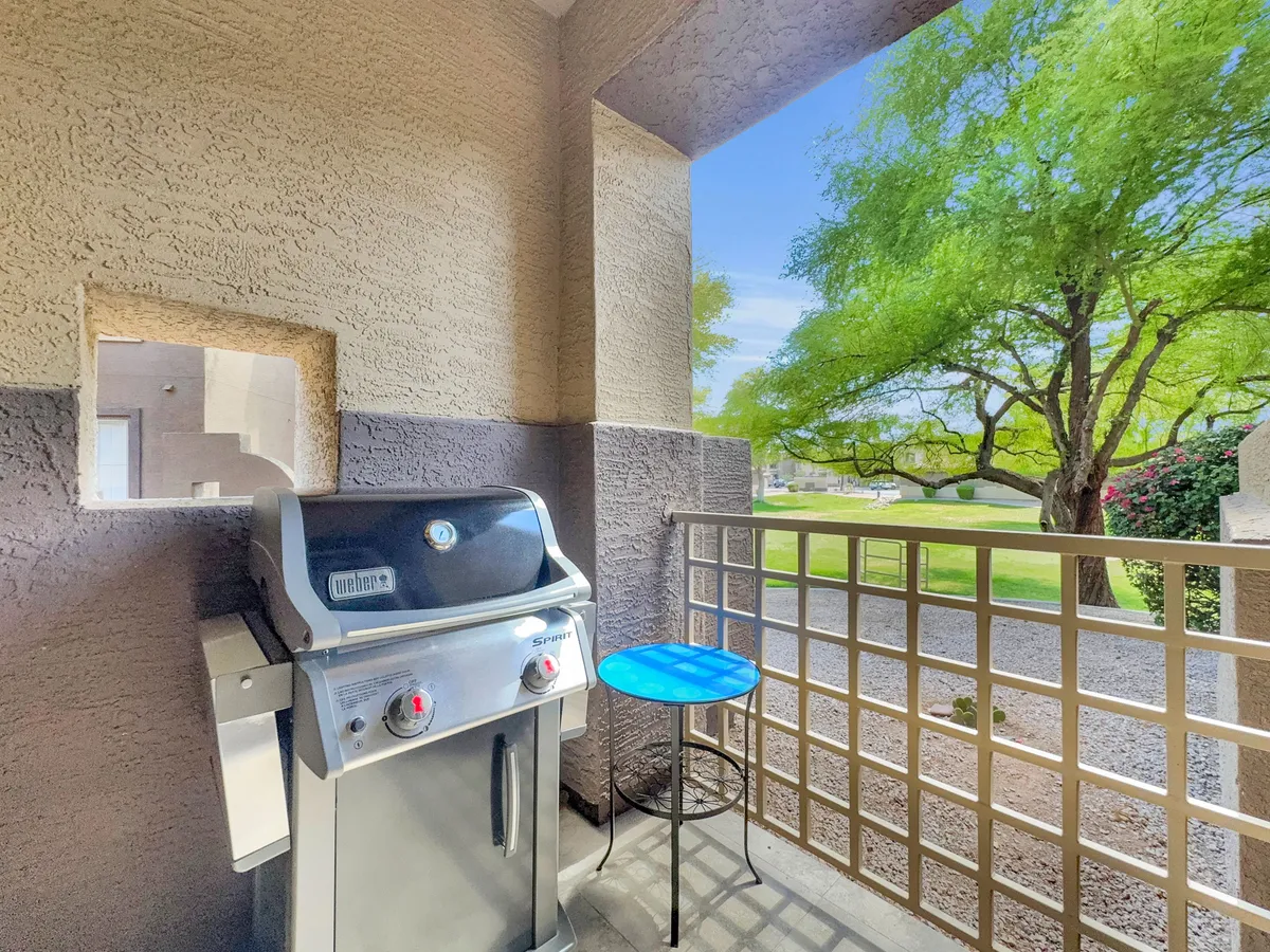 The image depicts an outdoor area featuring a grill, a small blue table, and a leafy green tree, with views extending into the surroundings. The scene suggests a relaxed setting for barbecuing or enjoying meals outdoors, with plenty of space for socializing.
