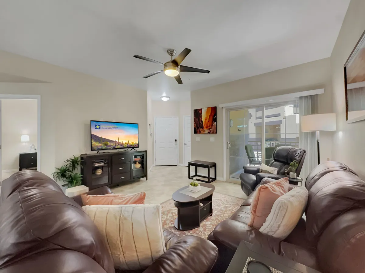 A living room scene featuring leather sofas arranged around a coffee table, with a TV mounted on the wall displaying scenic imagery. Bright natural light enters through sliding glass doors, hinting at outdoor amenities and communal gathering opportunities. Artwork adds a touch of personality to the welcoming space.